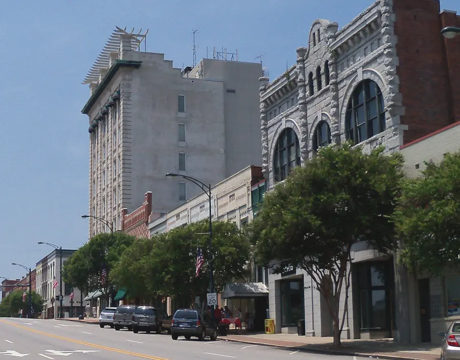 A wide street in downtown Salisbury, North Carolina, lined with historic brick and stone buildings, a tall white hotel block and flowering crape myrtles under a blue sky